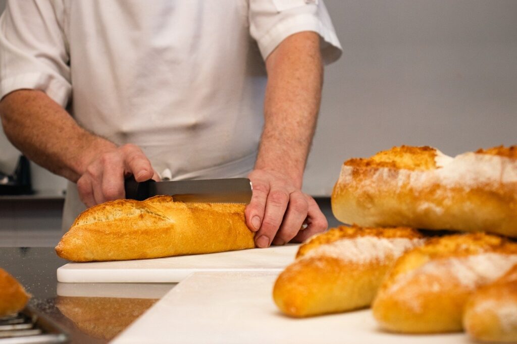 Un boulanger en tenue de travail coupe du pain sur une planche à découper dans une boulangerie.